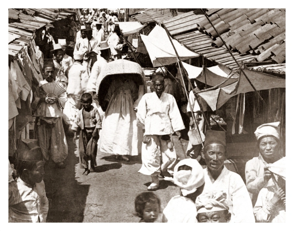 korean-girl-hides-under-a-large-basket-while-walking-down-a-crowded-side-street-in-1904-pyongy...png