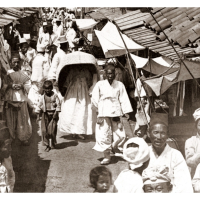 korean-girl-hides-under-a-large-basket-while-walking-down-a-crowded-side-street-in-1904-pyongy...png